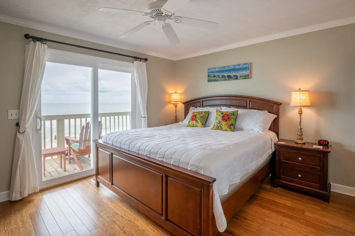 A serene primary bedroom with a king-size bed faces a sunny window, accented by decorative pillows and a stylish nightstand with a warm lamp. Soft, neutral tones and an inviting atmosphere create a tranquil retreat, and the window hints at possible deck access for a seamless indoor-outdoor experience.