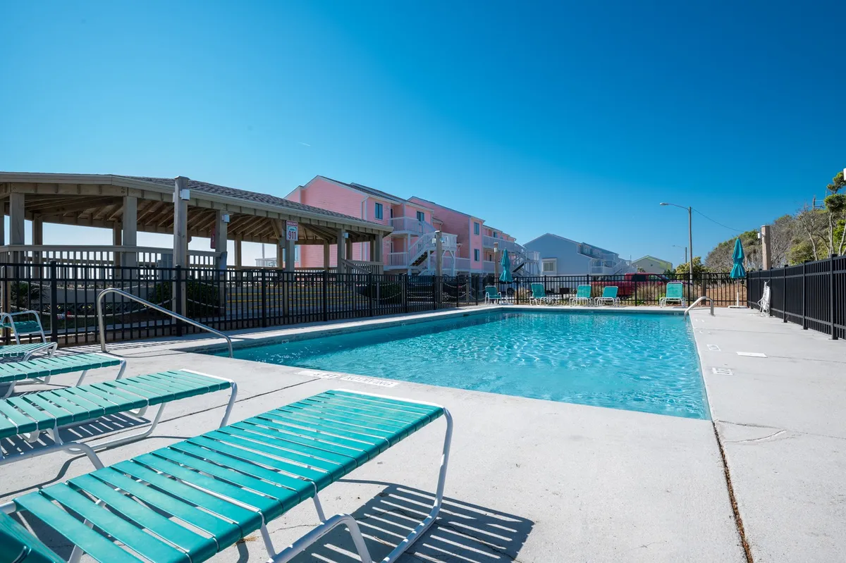A lively outdoor swimming pool area surrounded by colorful resort buildings and numerous lounge chairs. Guests are enjoying the pool, sunbathing, and socializing, creating an energetic, family-friendly atmosphere. The scene is lit by a warm sunset, adding to the vacation vibe.