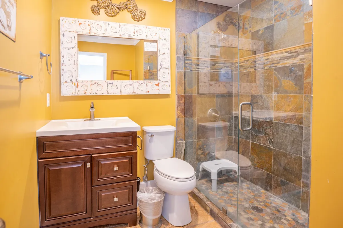 A modern ensuite bathroom featuring a spacious glass shower enclosure, sleek modern sink with a large decorative mirror, and a textured stone accent wall. The calming tones and spa-inspired design create an environment ideal for rejuvenation after a day by the coast.