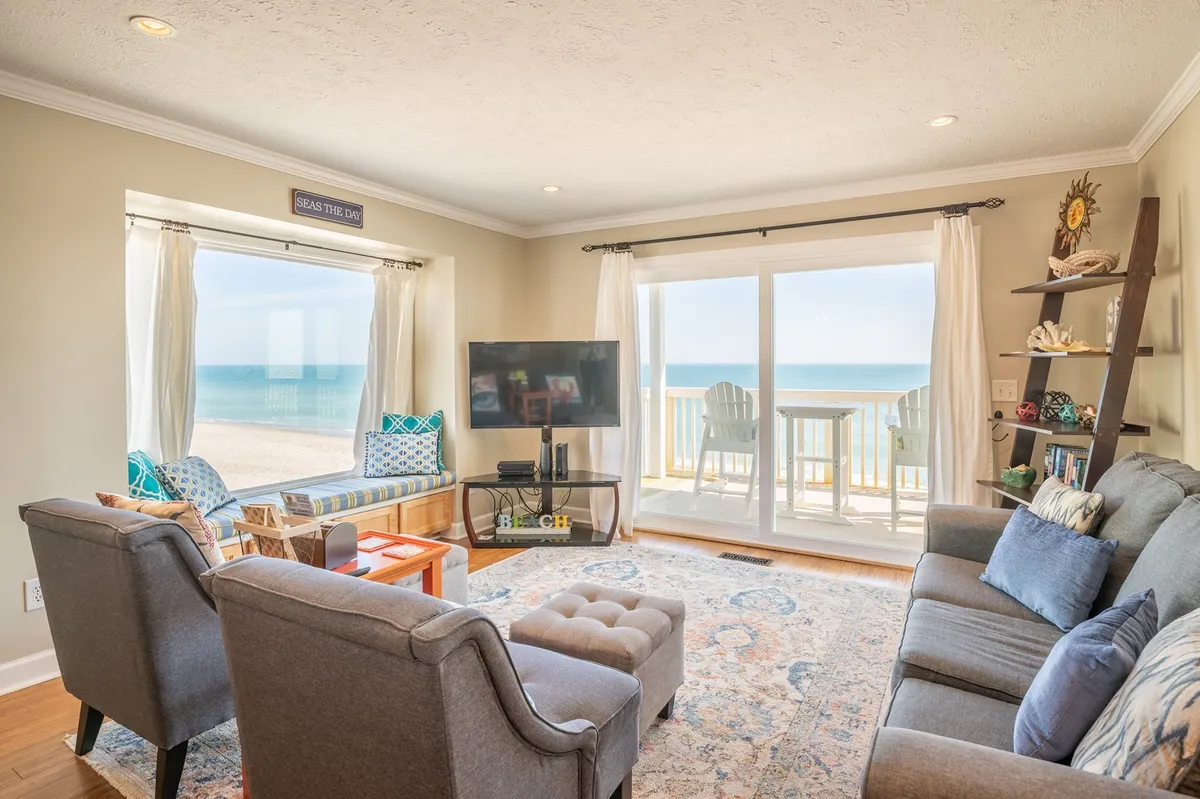The image showcases a bright, inviting living room with large windows framing sweeping ocean views. Comfortable sofas and stylish décor create a welcoming atmosphere for relaxation, while a television and decorative shelves add to the home's cozy charm. The scene exudes a sense of calm and togetherness, perfect for unwinding after a sun-soaked day at the beach.