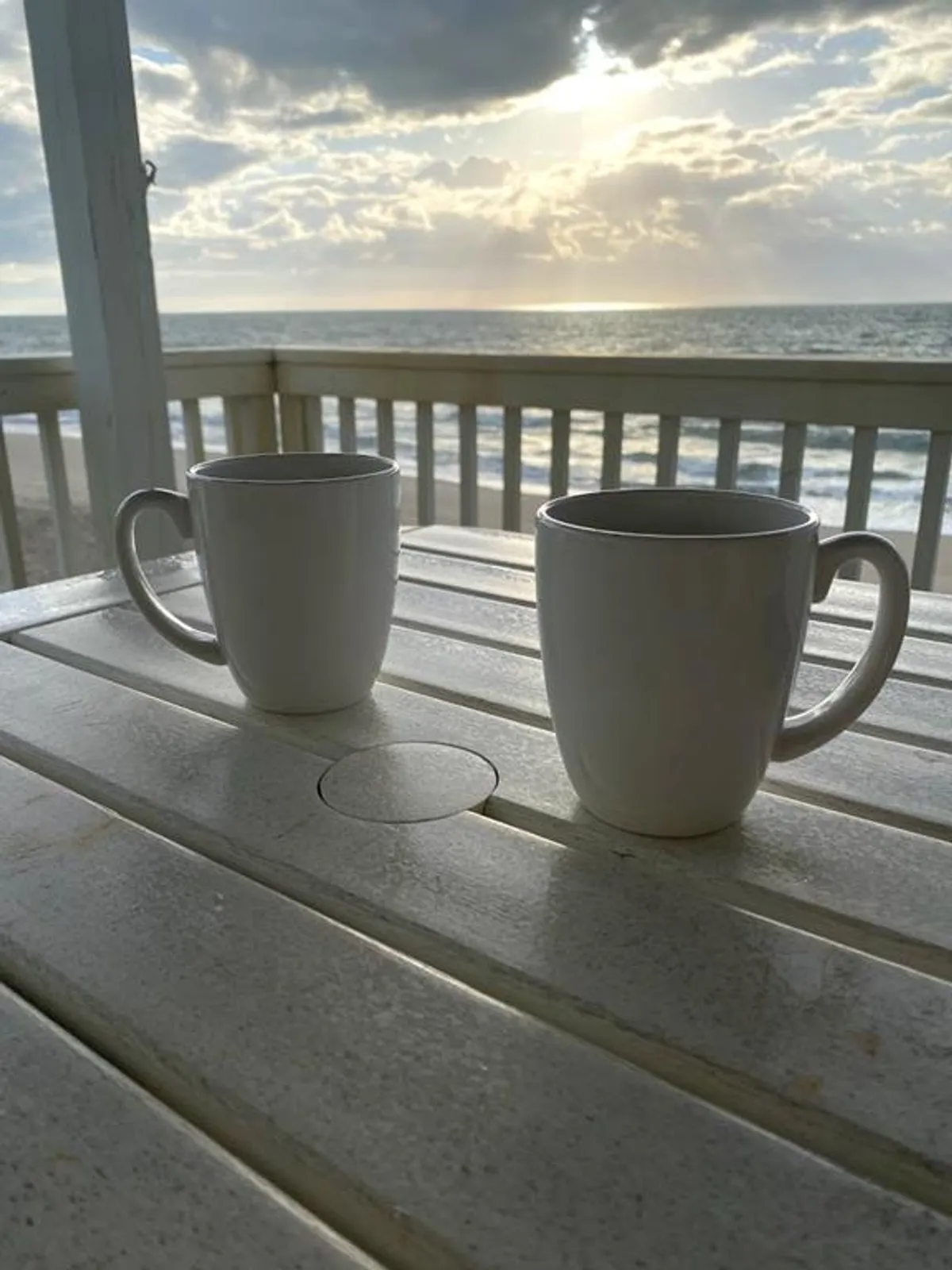 This image captures two white coffee mugs on a wooden table set on a balcony, overlooking a tranquil ocean view under soft sunlight and clouds. The setting feels serene and intimate, perfect for quiet mornings or romantic sunset moments.
