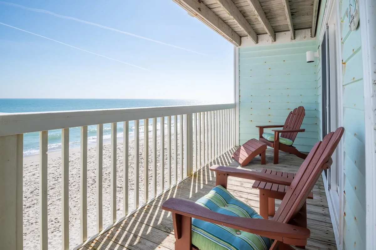 Two Adirondack chairs on a deck overlook a wide expanse of sandy beach and rolling ocean waves. The scene conveys peaceful relaxation with an immersive view of the coastline, ideal for watching wildlife or enjoying the ocean breeze.