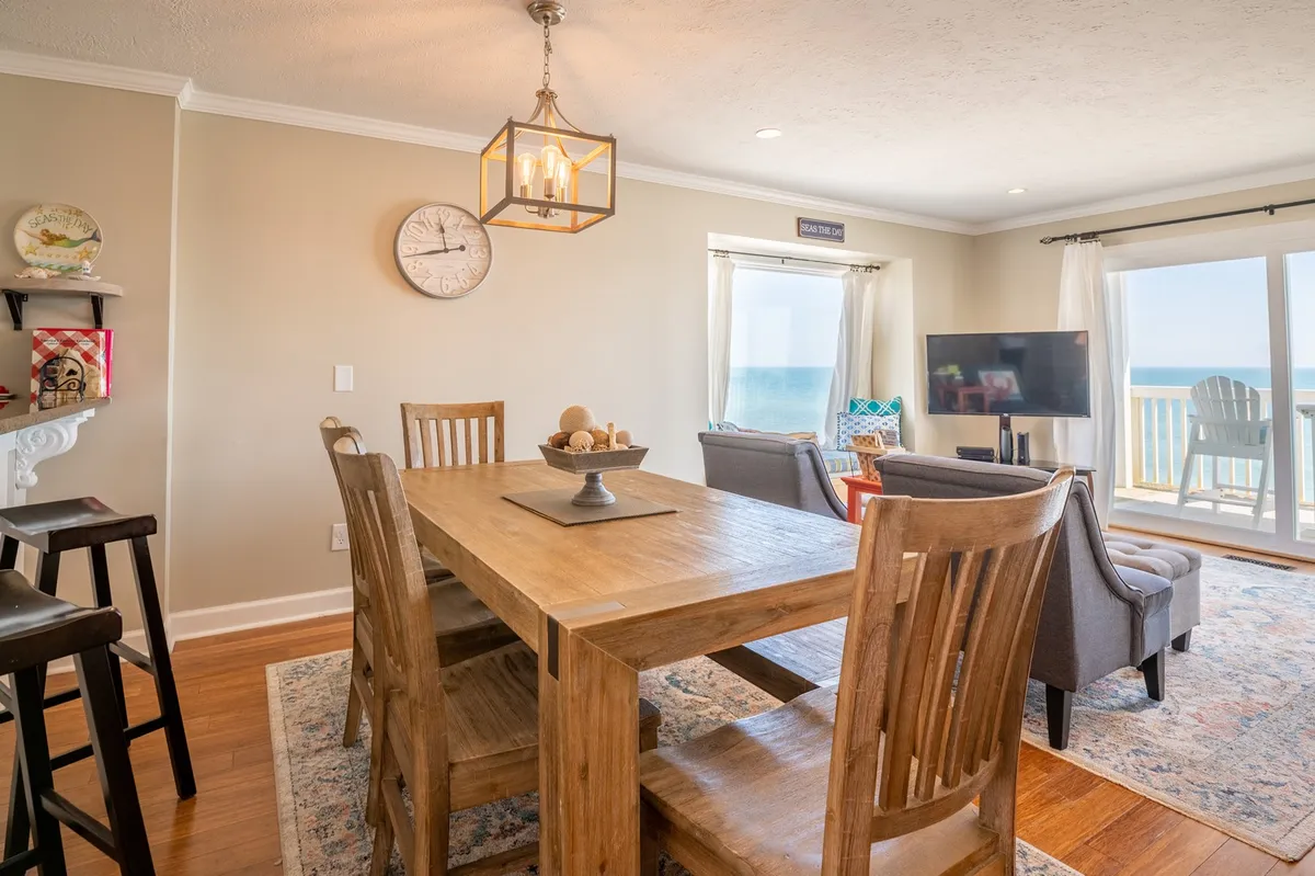 A dining area with a large table positioned near a window offering a stunning ocean view. The space is warmly lit and includes a television, blending mealtime with opportunities for game nights or movie watching. The inviting decor and layout encourage group gatherings and shared experiences.