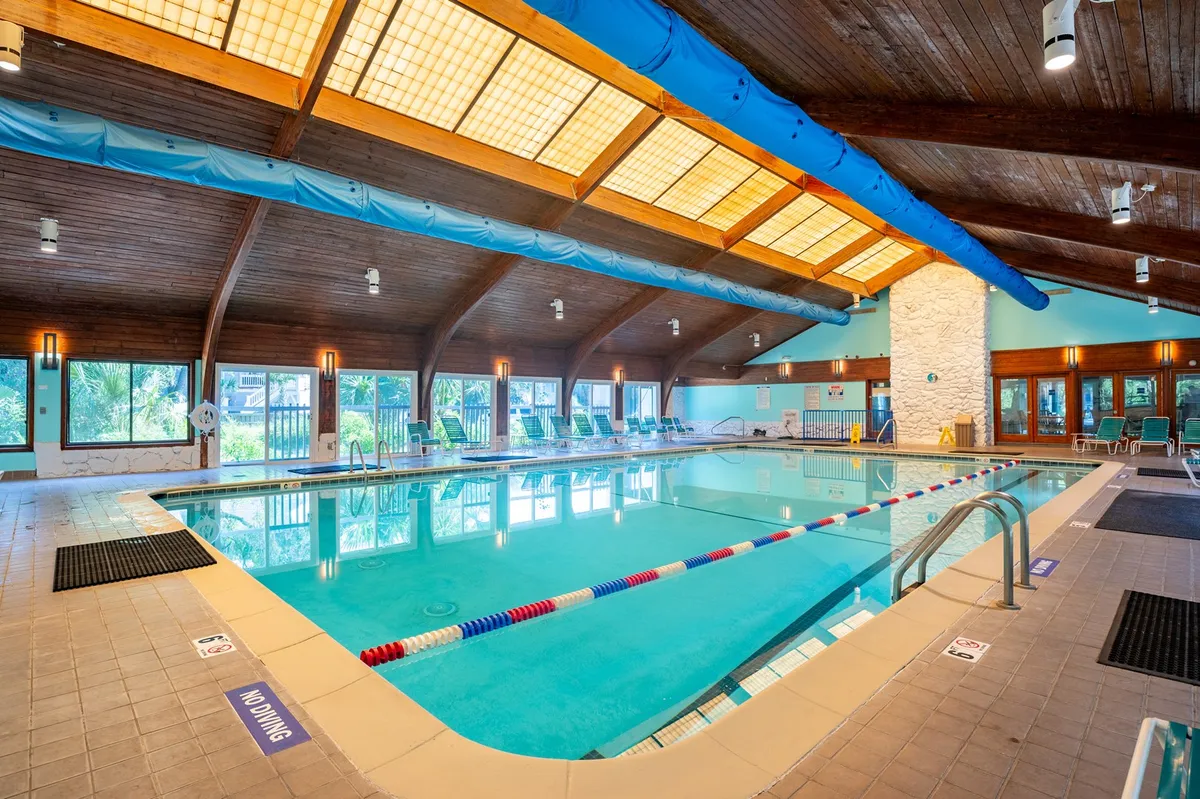 A bright, inviting indoor recreation center with a large swimming pool, wooden beams, and expansive windows that fill the space with natural light. Swimming lane markers are visible, and loungers line the side, suggesting both active and relaxing uses.