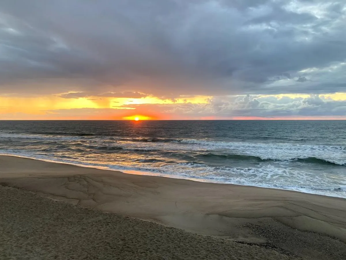 A panoramic view of a sandy beach with glistening ocean waves stretching toward the horizon, illuminated by the rich hues of a setting sun. The sky is streaked with warm colors, and the shoreline appears tranquil and uncrowded, emphasizing natural beauty and serene seclusion.
