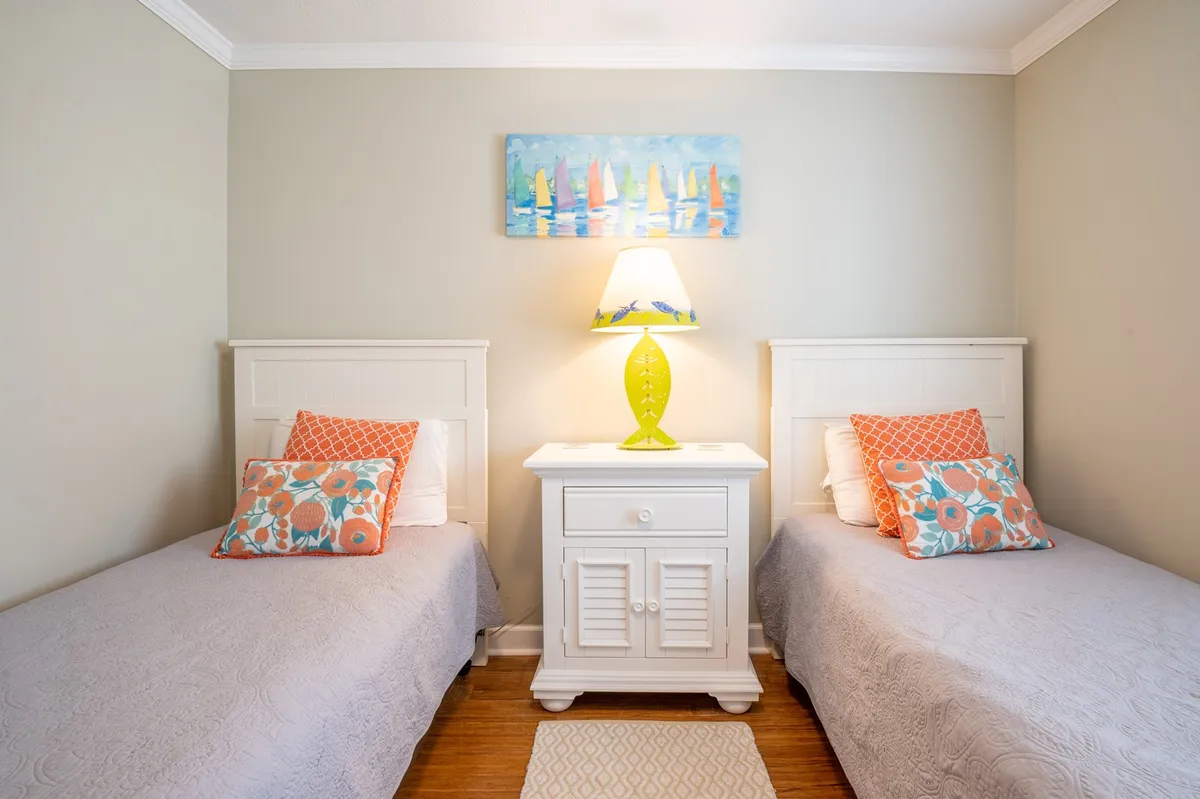 A cheerful guest bedroom with two twin beds, each adorned with colorful pillows and coordinated bedding. Above the beds hangs playful artwork, and a bright lamp sits between them on a nightstand. The mood is vibrant yet restful, ideal for kids or friends.
