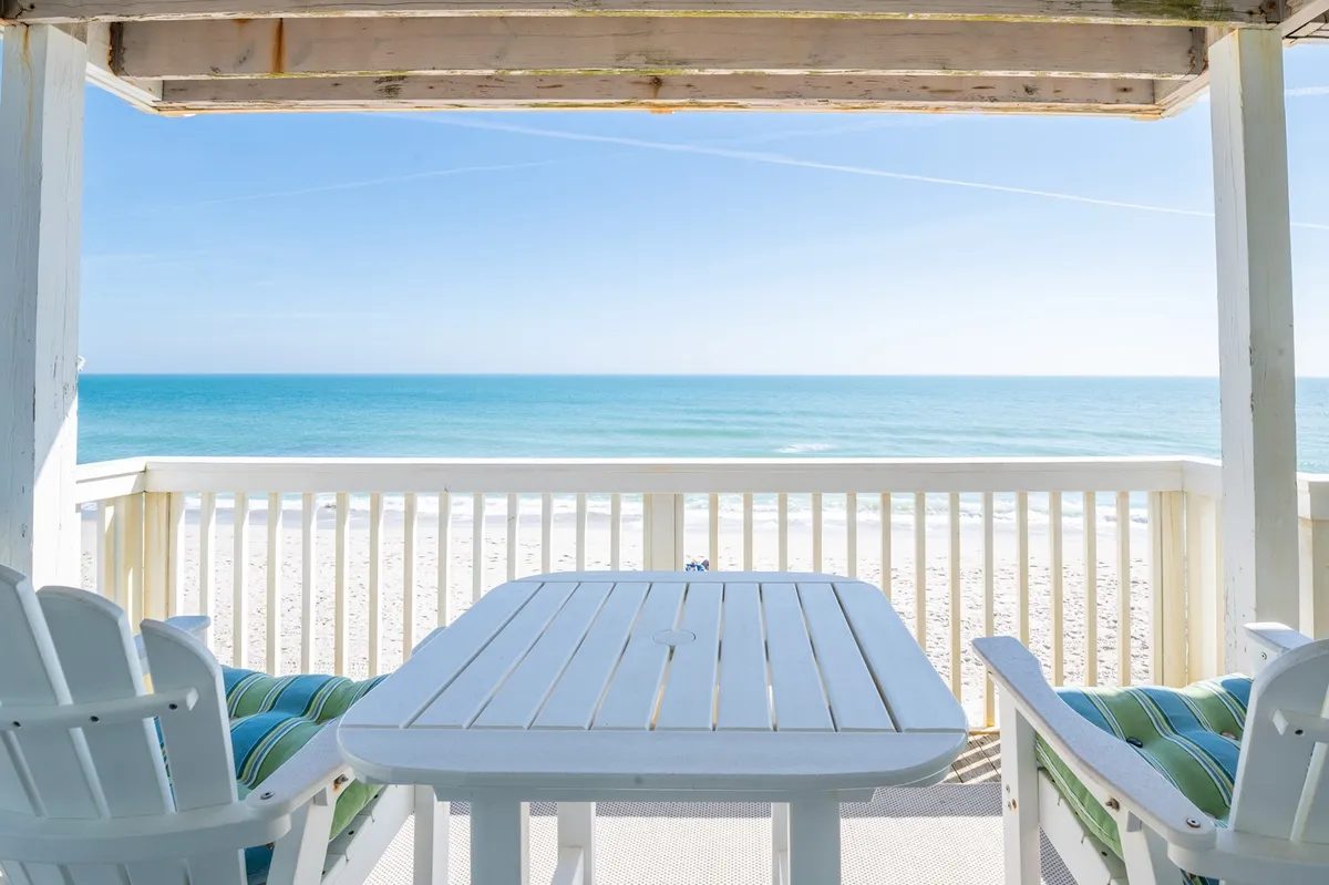 An oceanfront wooden deck with comfortable outdoor furniture, overlooking expansive water views. The deck invites relaxation and socializing, seamlessly connecting the indoor space to the natural beauty outside. Sunlight dances on the water, creating a tranquil setting for gatherings or quiet moments.