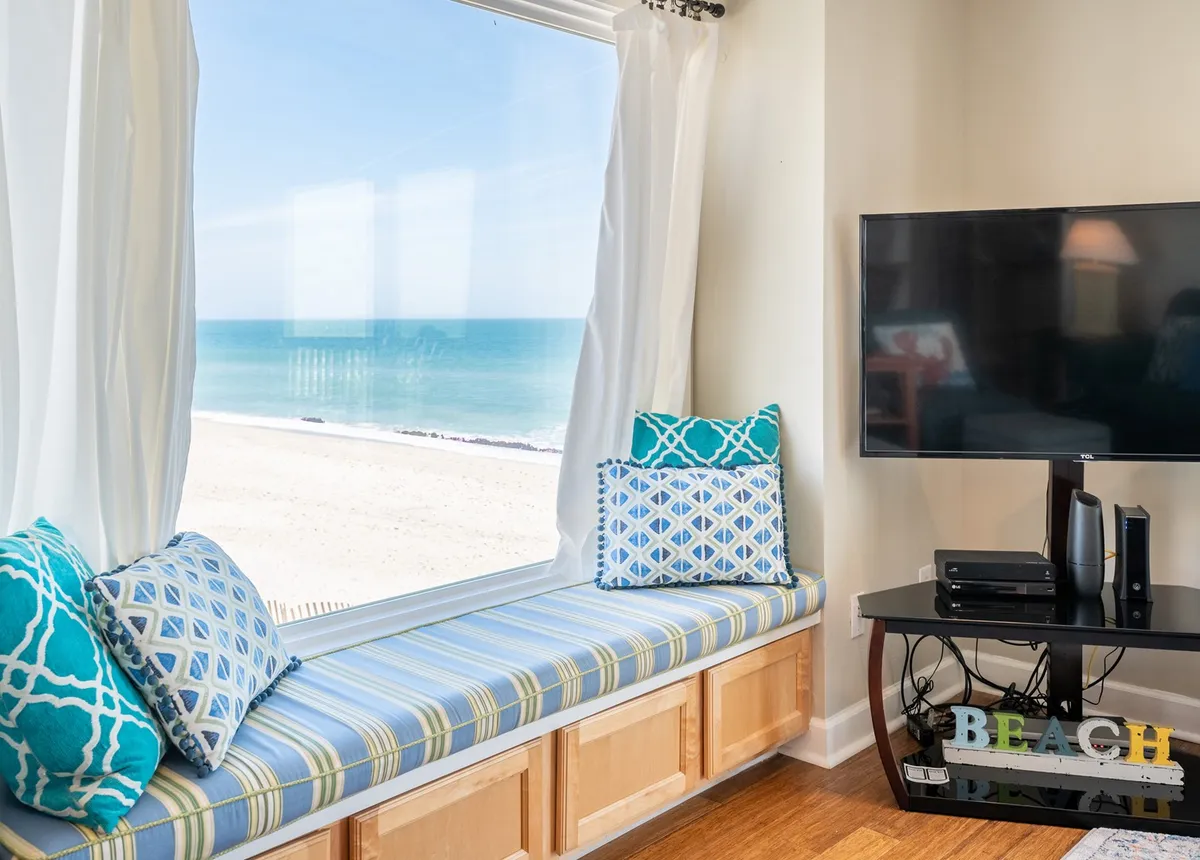 A sunlit living room with a built-in window seat, plush cushions, and panoramic ocean views through large windows. A television is visible, making the space inviting for movie nights and family gatherings. The serene atmosphere is enhanced by the natural sunlight pouring in, highlighting both comfort and scenic beauty.